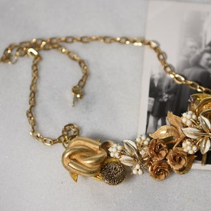 Può includere: Una collana d'oro con un grande ciondolo decorato con un gruppo di fiori, foglie e perle finte. Il ciondolo è attaccato a una catena d'oro.