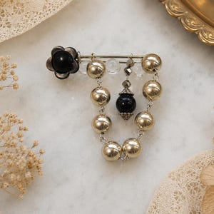 Peut inclure: Broche décorative avec une fleur noire et des perles dorées. La broche est dotée d'une épingle et d'une chaîne de perles pendantes. Des perles transparentes et noires ajoutent des détails, créant un accessoire élégant.