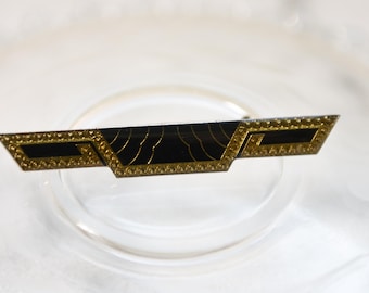 Könnte beinhalten: Eine schwarze und goldene Brosche mit geometrischem Design. Die Brosche hat eine schwarze Emaille-Mitte mit goldenen Akzenten und einem goldenen Rand.