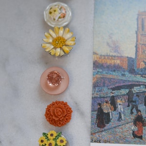 May include: A selection of decorative buttons with floral designs and a postcard featuring an impressionist painting. The buttons include a yellow daisy, a pink button with a crystal, and a button with a terracotta rose.