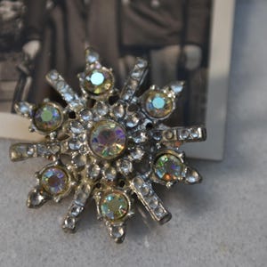 May include: A silver-toned brooch with a starburst design. The brooch is adorned with iridescent rhinestones.