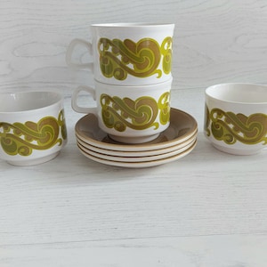 May include: Set of four white teacups with a green and brown swirl pattern. The cups are stacked on top of each other and are sitting on a white wooden surface.