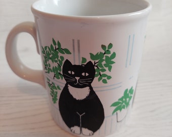 May include: White ceramic mug with a black cat and green plants design. The cat is sitting in front of a window with blue and white stripes.