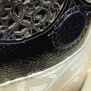 Puede incluir: Primer plano de un objeto negro y ornamentado, posiblemente de cuero, con una superficie texturizada y un acento ovalado. El objeto tiene un dise&ntilde;o decorativo en relieve con un acabado dorado met&aacute;lico. Una tela color crema es visible en la parte inferior de la imagen.