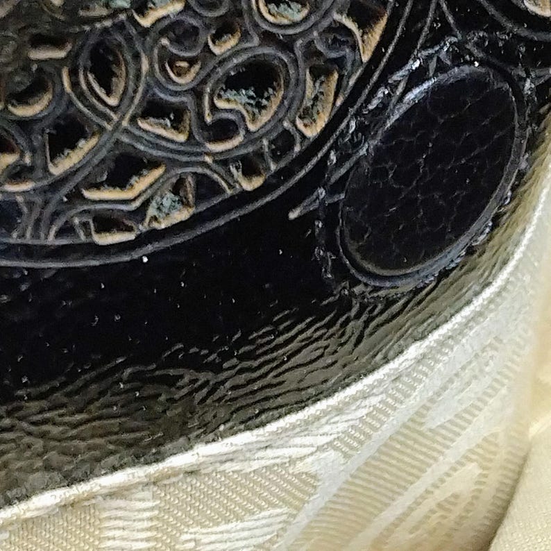 Puede incluir: Primer plano de un objeto negro y ornamentado, posiblemente de cuero, con una superficie texturizada y un acento ovalado. El objeto tiene un dise&ntilde;o decorativo en relieve con un acabado dorado met&aacute;lico. Una tela color crema es visible en la parte inferior de la imagen.