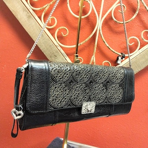 Puede incluir: Bolso de mano de cuero negro con una cadena plateada y un cierre decorativo plateado. La parte delantera del bolso presenta un dise&ntilde;o de filigrana plateada. Un dije de coraz&oacute;n plateado cuelga del lateral. El bolso se exhibe sobre un fondo rojo.