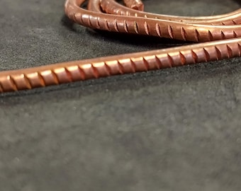 May include: A coiled length of metallic copper wire featuring a repeating pattern of small, evenly spaced vertical indentations along its entire surface. The wire has a warm, reddish-brown metallic finish and is arranged in a circular loop.