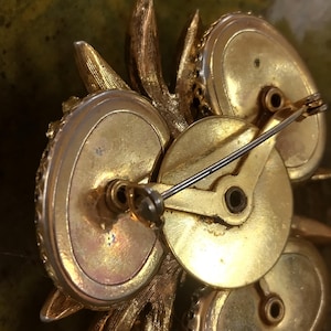 May include: The reverse side of a gold-toned metal brooch featuring a textured leaf-like design. The back displays a circular central plate with a hinged pin mechanism and a rotating safety clasp, showing visible signs of age and metallic patina.