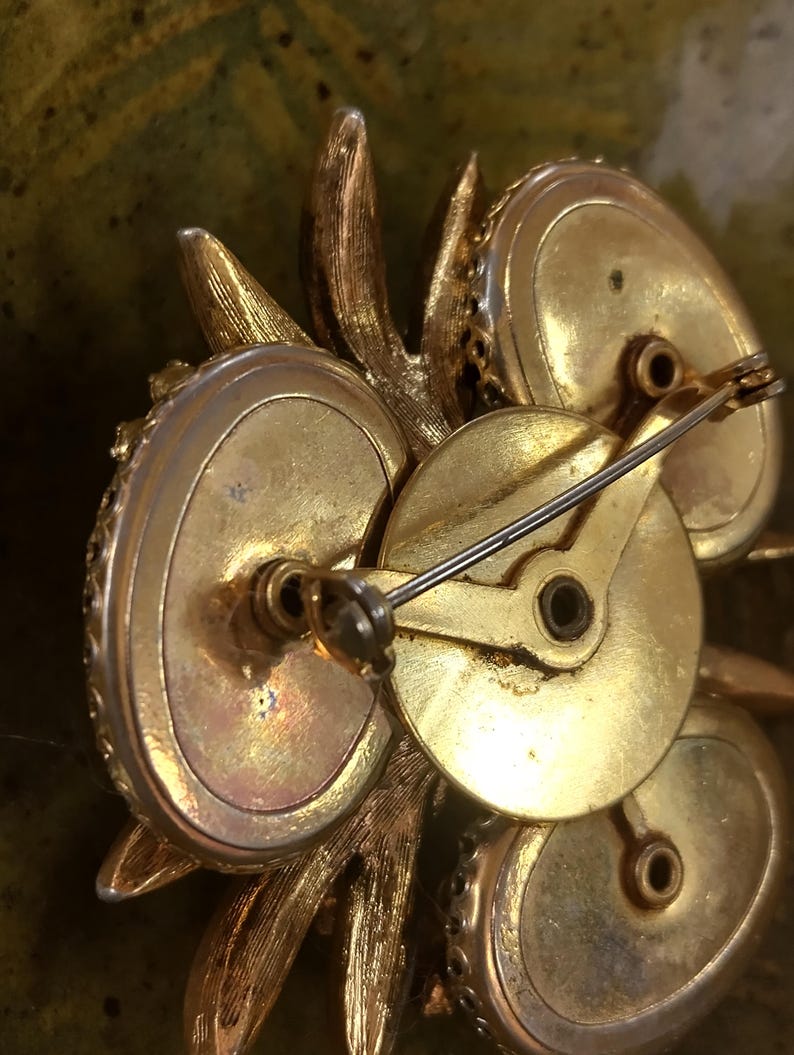May include: The reverse side of a gold-toned metal brooch featuring a textured leaf-like design. The back displays a circular central plate with a hinged pin mechanism and a rotating safety clasp, showing visible signs of age and metallic patina.