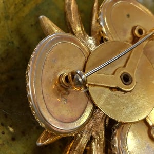 May include: A gold-toned metal brooch viewed from the back, featuring a pin mechanism with a rotating safety clasp. The structure consists of multiple circular settings and textured leaf-like elements, with a visible maker's mark on the metal surface.