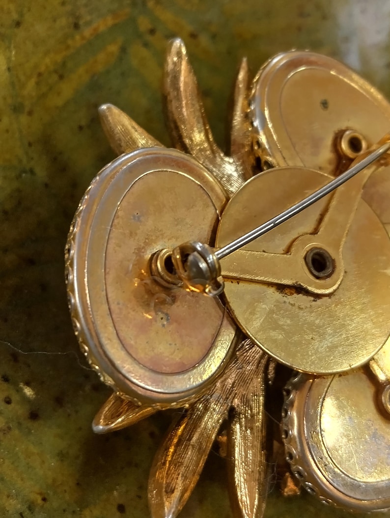 May include: A gold-toned metal brooch viewed from the back, featuring a pin mechanism with a rotating safety clasp. The structure consists of multiple circular settings and textured leaf-like elements, with a visible maker's mark on the metal surface.