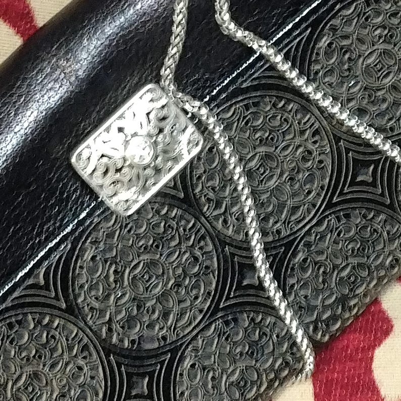 Puede incluir: Collar con colgante de filigrana plateada con un colgante rectangular. El colgante presenta un intrincado dise&ntilde;o calado y una peque&ntilde;a piedra transparente en el centro. La cadena del collar es de estilo trenzado delicado. El colgante descansa sobre una superficie de cuero negro.