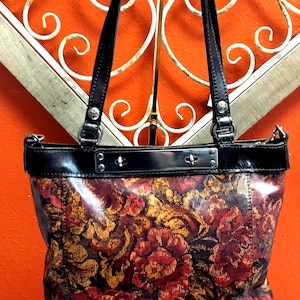May include: A floral-patterned tote bag with black leather handles and trim. The bag features a vibrant design of red and gold flowers on a dark background. The handles are attached with silver-toned hardware.