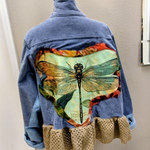 May include: A blue denim jacket with a large dragonfly design on the back. The dragonfly is in shades of green, orange, and brown. The jacket has a crochet trim at the bottom and rolled-up sleeves.