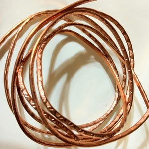 May include: Coiled copper wire with a textured surface. The wire is a warm, reddish-brown color and appears to be handmade. The wire is arranged in a loose, circular pattern against a white background.
