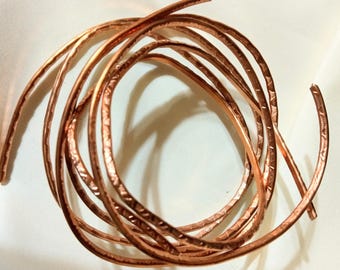 May include: Coiled copper wire with a textured surface. The wire is a warm, reddish-brown color and appears to be handmade. The wire is arranged in a loose, circular pattern against a white background.