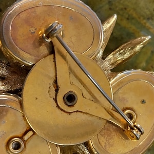 May include: A gold-tone metal brooch featuring a cluster of three oval-shaped components arranged around a central circular plate. The back includes a pin mechanism with a rotating clasp, showing textured leaf-like accents extending from the sides.