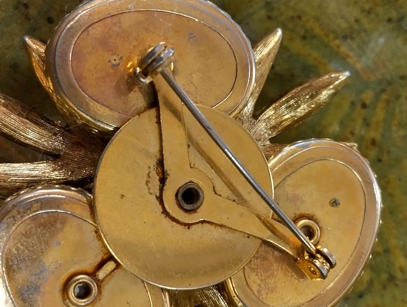 May include: A gold-tone metal brooch featuring a cluster of three oval-shaped components arranged around a central circular plate. The back includes a pin mechanism with a rotating clasp, showing textured leaf-like accents extending from the sides.