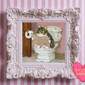 May include: A framed painting depicts a cat reading a book titled "Cat News" while sitting on a toilet. The painting is in a white ornate frame against a pink and white striped background. A pink heart with the text "KITSCHY DARLING" is in the bottom right corner.