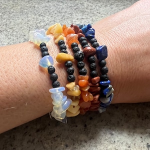 May include: A collection of five gemstone bracelets in a rainbow of colors. Each bracelet features a strand of black lava beads and a variety of colorful, irregularly shaped gemstones. The bracelets are stacked on a wrist.