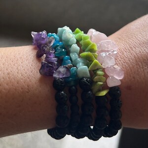 May include: A collection of gemstone bracelets in various colors, including purple, blue, green, and pink. The bracelets are made with rough-cut stones and black lava beads. The bracelets are stacked on a wrist.