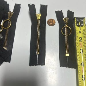 May include: Close-up of three black zippers with gold-colored metal teeth and pulls. One zipper has a circular pull, another has a rectangular pull, and the third has a standard pull. A penny and a tape measure are also visible.