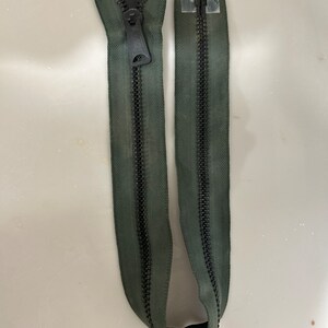 Puede incluir: Una cremallera verde con un deslizador y dientes negros. La cremallera está separada en dos mitades.