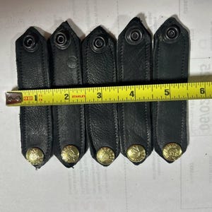 Puede incluir: Seis correas de cuero negro con botones de metal dorado. Las correas están alineadas junto a una cinta métrica amarilla.