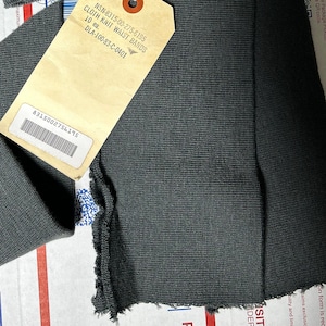 Könnte beinhalten: Nahaufnahme eines dunkelgrauen Strickstoffes mit einem daran befestigten Etikett. Das Etikett ist beige und trägt folgenden Text: NSN-8315-00-275-6195 CLOTH KNIT WAIST BANDS 10 ea. DLA-100-83-C-0401. Das Etikett hat auch einen Barcode mit der Nummer 8315002756195.