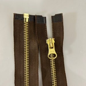 以下が含まれることがあります： 金色の金属製の歯と引き手が付いた、3つの茶色のジッパーのクローズアップ。ジッパーの引き手には「IDEAL」の文字が刻まれています。ジッパーは分離されており、歯と布テープが表示されています。