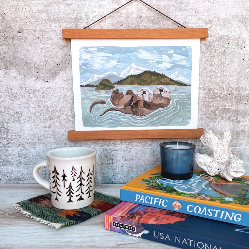 May include: A framed illustration of two sea otters floating on their backs in the water. The otters are brown and white and have their paws in the air. The background is a blue sky with white clouds and a mountain range in the distance.