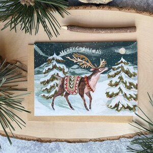 May include: A Christmas card featuring a reindeer in a snowy landscape. The reindeer has large antlers and is wearing a decorative blanket. The card reads "MERRY CHRISTMAS" in gold lettering against a night sky with a full moon.