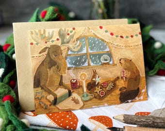 May include: A Christmas card depicting a moose and a beaver baking cookies in a cosy cabin. The moose is rolling out dough with a rolling pin, while the beaver is decorating cookies with red and white icing. The scene is set against a snowy window with a snow globe on the sill.