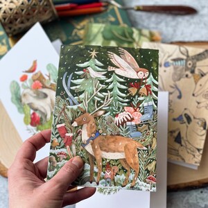 May include: A Christmas card featuring a watercolor illustration of a deer in a snowy forest with a white owl, a Christmas tree, and a red and white candy cane. The card has a green background with white snowflakes.