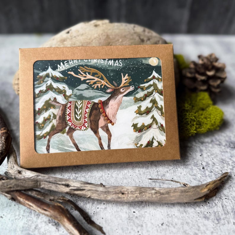 May include: A Christmas card featuring a watercolor illustration of a reindeer in a snowy forest. The reindeer has large antlers and is wearing a decorative blanket. The words "MERRY CHRISTMAS" are written at the top of the card.