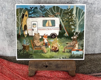 May include: A whimsical illustration depicts a camping scene with a white camper van and various forest animals. A bear, fox, and deer are gathered around a campfire roasting marshmallows. The word "CAMP" is printed on the side of the camper.