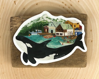 May include: A sticker featuring a black and white orca whale swimming over a colorful illustration of a small town with houses and boats. The text "Canyon & Cove" is printed on the whale's body.