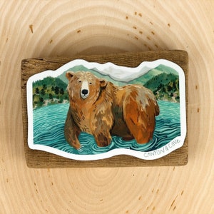 Op de afbeelding: Een sticker met een bruine beer die in een meer staat met groene bomen en bergen op de achtergrond. De sticker heeft een golvende rand en staat onderaan "Canyon & Cove".