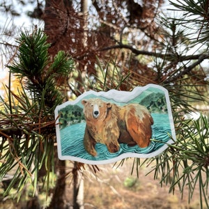 Op de afbeelding: Een sticker met een bruine beer die in een blauw meer staat met groene bomen op de achtergrond.