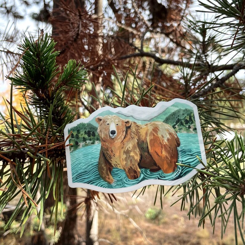 Op de afbeelding: Een sticker met een bruine beer die in een blauw meer staat met groene bomen op de achtergrond.