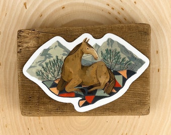 May include: A sticker featuring a brown horse lying down on a colourful patchwork quilt. The horse is in a mountainous landscape with trees and the text "Canyon Cove" is visible in the top right corner.
