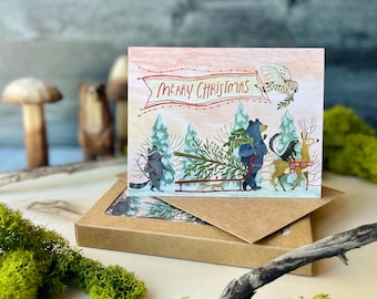 Pode incluir: Um cartão de Natal com um grupo de animais da floresta puxando uma árvore de Natal em um trenó. O cartão tem uma faixa vermelha com o texto "Feliz Natal" e um fundo de floresta com neve.