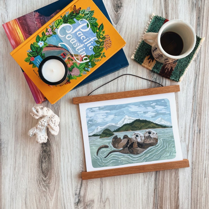 May include: A framed print of two sea otters floating on their backs in a blue ocean with mountains in the background. The print is in a wooden frame with a black hanging wire.