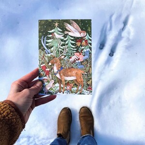 May include: A Christmas card featuring a reindeer with a red and white bell collar standing in a snowy forest. The reindeer is surrounded by trees, a white owl, and other woodland creatures. The card has a green background with a festive design.