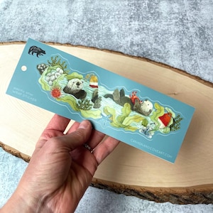 May include: A blue and green sticker with a sea otter floating on its back in the water. The otter is holding a red crab. The sticker has the text "Sea Otter Wrap Sticker" and "canyonandcoveart.com".