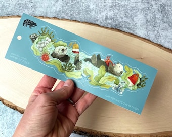 May include: A blue and green sticker with a sea otter floating on its back in the water. The otter is holding a red crab. The sticker has the text "Sea Otter Wrap Sticker" and "canyonandcoveart.com".