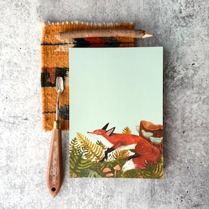 May include: A light green notepad with a watercolor illustration of a red fox resting in a forest setting. A wooden pencil, a small palette knife, and a piece of patterned fabric are arranged around the notepad.