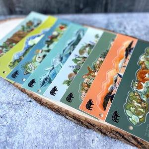 May include: A set of eight colorful bookmarks featuring different animal illustrations. Each bookmark has a black bear silhouette and the text "Wild Things" at the bottom. The bookmarks are arranged in a row on a wooden surface.