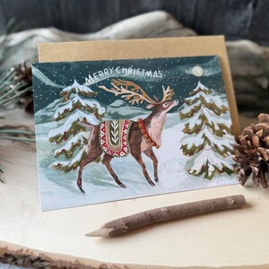 May include: A Christmas card featuring a reindeer in a snowy forest scene. The reindeer has large antlers and is wearing a decorative blanket. The card has the words "MERRY CHRISTMAS" at the top. A brown envelope is behind the card.