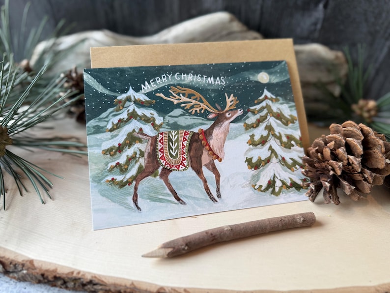 May include: A Christmas card featuring a reindeer in a snowy forest scene. The reindeer has large antlers and is wearing a decorative blanket. The card has the words "MERRY CHRISTMAS" at the top. A brown envelope is behind the card.
